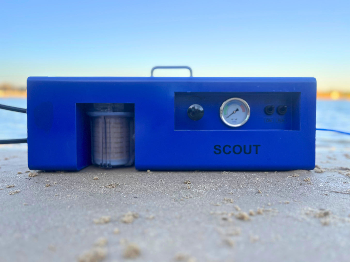 Blue device labeled 'SCOUT' on a sandy surface with a blurred natural background
